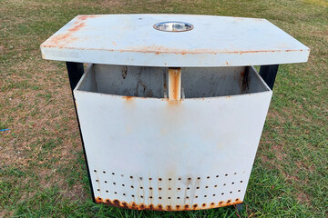 A white outdoor trash bin made of metal, showing signs of rust and weathering, placed on a grassy area.