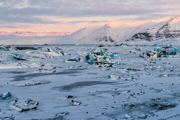 Icebergs and huge chunks for green-blueish ice fill Jokulsarlon Glacier lagoon, as the golden hour sunlight of the early morning casts a beautiful warm glow over the glacier-filled national park in