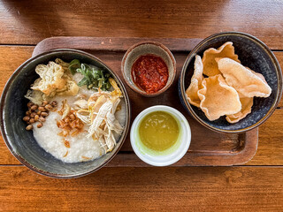 Traditional Indonesian chicken porridge or Bubur Ayam served with crackers, chili sauce, green sauce, and toppings on a wooden tray over rustic wooden table.