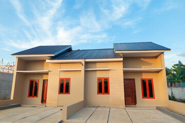 A simple permanent house with a modern minimalist design, beige walls, wooden doors, and red-framed windows, viewed from the front under a clear blue sky.