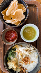 Traditional Indonesian chicken porridge or Bubur Ayam served with crackers, chili sauce, green sauce, and toppings on a wooden tray over rustic wooden table.