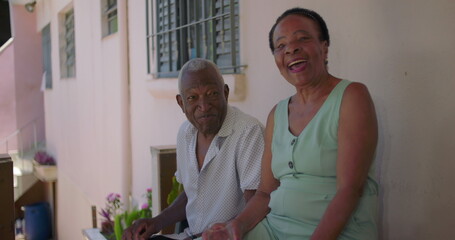African American older couple enjoying a lighthearted moment while sitting outside at residence, the woman smiling joyfully while holding a glass with a drink, raising it in a cheerful toast