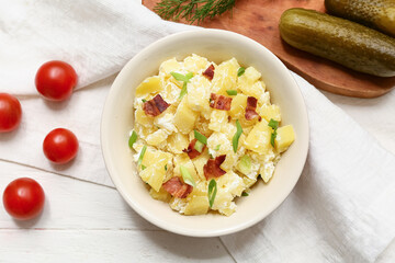 Bowl of tasty potato salad with bacon, green onion, tomatoes and pickled cucumbers on white wooden background