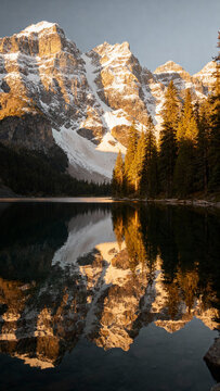 Snowy mountains reflected in a clear alpine lake at golden hour, serene nature scene with pines and soft light.Generated by AI