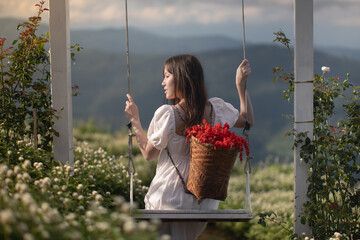 A young Asian woman in a white dress playing on the swing in a field of white amaranthus flowers surrounded by mountains and mist