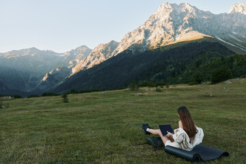 Fototapeta premium A solitary worker sits on a mat in a wide meadow, laptop in lap, while towering mountains rise nearby, creating a calm, productive outdoor work scene.