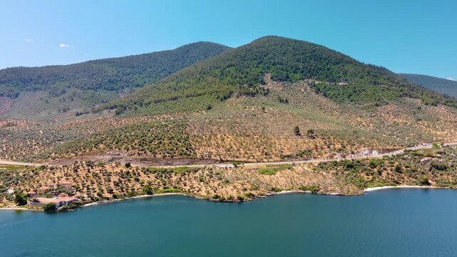 aerial footage showcasing the tranquil blue waters of Bafa Lake, nestled amidst the rugged, tree covered mountains of Turkey. This serene natural landscape offers a glimpse into the beauty of the Turk