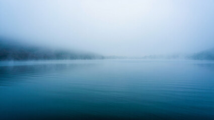 Calm lake surface reflecting the sky and trees obscured by thick fog creating a serene landscape