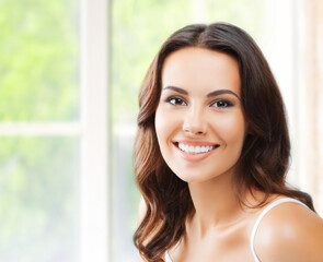 Portrait image of young cute dark brunette wavy hair woman cheerful smiling confidently at camera, set against window background with blurred greenery, home