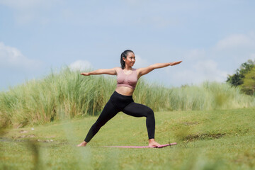 Fototapeta premium Mental and physical health. A slender, tanned Asian girl in sportswear does yoga by a calm river in the morning. The woman stands in a warrior pose with her arms outstretched.