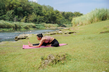 Mental and physical health. A slender, tanned Asian woman in sportswear performs yoga on a calm riverbank in the morning. The woman is performing Janu Sirsasana, or Head-to-Knee Forward Bend.