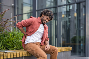 A young Indian man sits on a bench near a building, holding his back with his hand, feeling severe pain
