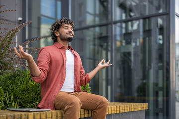Relaxed young Indian man sitting on a bench outside a building in lotus position with eyes closed and meditating