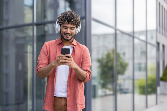 A young Indian man walks through the city wearing headphones and using a mobile phone
