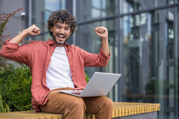 Portrait of a young Indian man sitting on a bench outside, holding a laptop on his lap and rejoicing in success with his arms raised