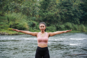 A slender, tanned Asian girl in sportswear doing arm stretching exercises in the open air on the edge of a beautiful river, before exercising.
