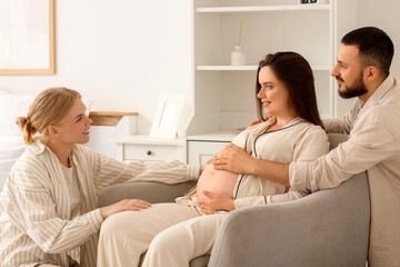 Obraz premium Young happy pregnant woman sitting on armchair with her husband and doula in bedroom