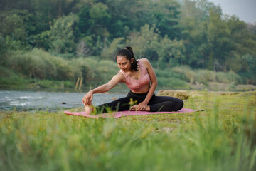 A slender Asian girl with brown skin in sportswear is doing stretching movements of her hamstrings in the open air on the edge of a beautiful river, before doing sports.
