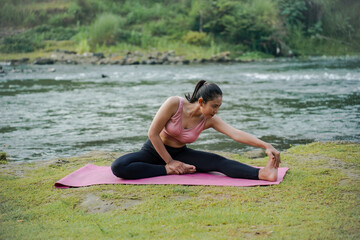 A slender Asian girl with brown skin in sportswear is doing stretching movements of her hamstrings in the open air on the edge of a beautiful river, before doing sports.
