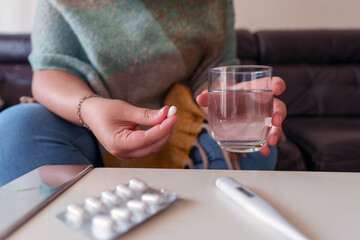 Woman holding a pill and water glass, taking medication for illness, surrounded by a thermometer and blister pack