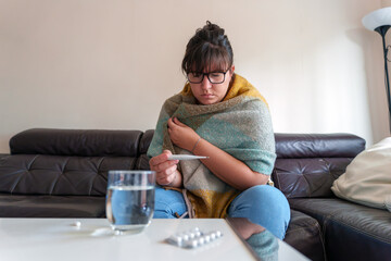 Obraz premium Woman checking temperature with a thermometer, feeling ill and experiencing flu symptoms at home