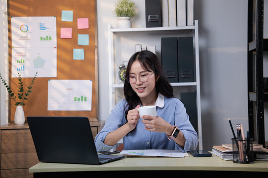 Young Asian woman wearing glasses, wearing a blue shirt, holding a cup of coffee sitting in an office room.