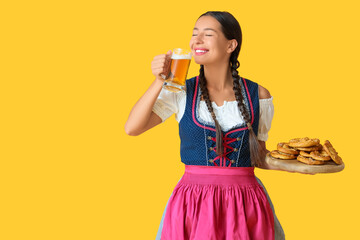 Young happy woman in traditional German clothes with glass of beer and many tasty pretzels on yellow background