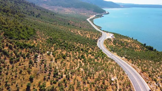 An aerial drone shot captures the serene beauty of Bafa Lake in Turkey, with a winding coastal road tracing its shoreline amidst lush, arid Mediterranean vegetation. The footage showcases the natural 