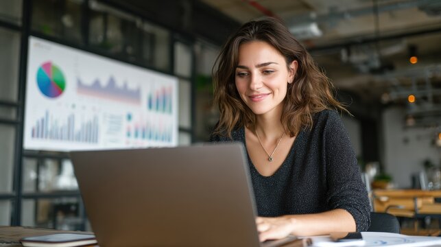 Focused woman working on a laptop, smiling in a modern office with data visualizations in the background.