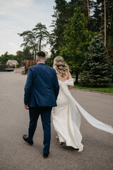 A bride in a white dress with a long veil walks through the park holding the hand of her groom in a classic suit, who is walking next to her