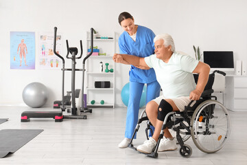 Female therapist helping senior man to stand up from wheelchair in rehabilitation center