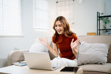 Obraz premium Cheerful Asian woman wearing headphones smiling and waving hands during online video call at home. Modern communication, friendship, and remote connection concept.