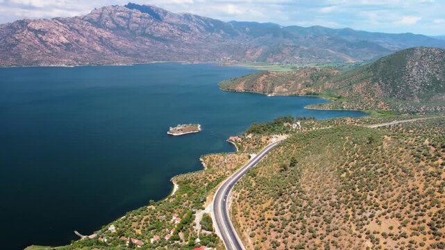 aerial drone shot capturing the serene beauty of Bafa Lake in Turkey, showcasing its deep blue waters, the surrounding lush green hills, and a winding coastal road. The footage highlights the natural 