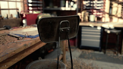 A worn metal chair stands in a cluttered workshop filled with tools and materials. Dust and shavings cover the floor, hinting at recent work. Light pours through the windows.