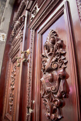Ornate colonial wooden door with floral carvings, antique emblem, and handcrafted fish-shaped door knockers, showcasing Mexican Baroque craftsmanship and historic elegance.