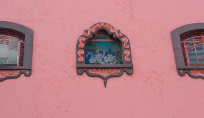 Colonial Mexican building with a pink facade and three symmetrical windows, featuring an antique leaded glass swan design and carved stone frames.