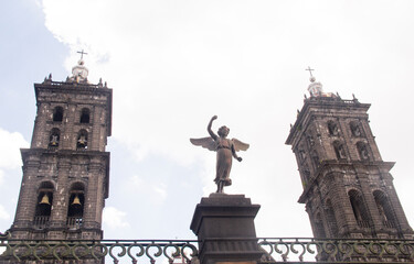 Detailed metal angel sculpture with torch at Puebla Cathedral, showcasing intricate craftsmanship with both iconic towers framing the historic Baroque architecture.