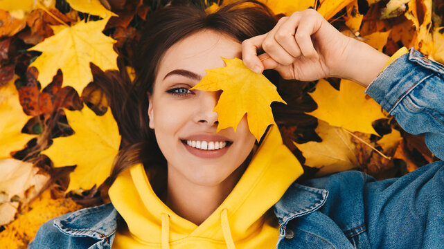 Smiling young woman with brown hair in yellow hoodie and denim jacket lying on autumn leaves, holding a maple leaf over her eye, joyful fall portrait.