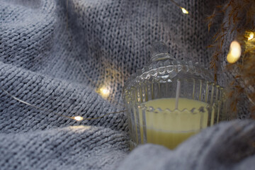 Close-up of candle in glass jar on gray knitted blanket. Knitted background. Cozy autumn or winter weekend.
