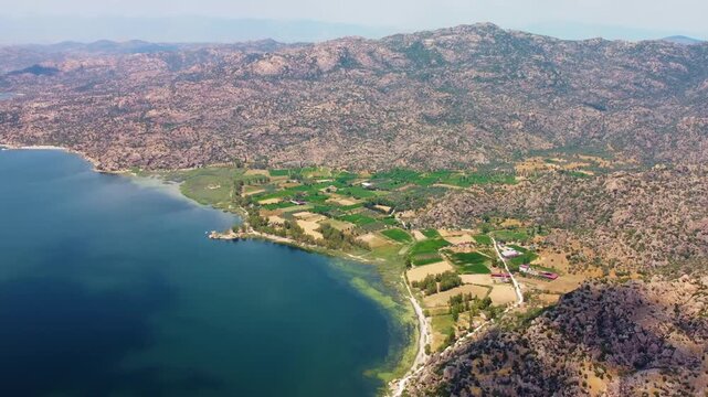 aerial perspective captures the serene beauty of Bafa Lake, a large freshwater lake in Turkey, nestled amidst rugged mountains and lush green agricultural fields. The footage showcases the stunning na