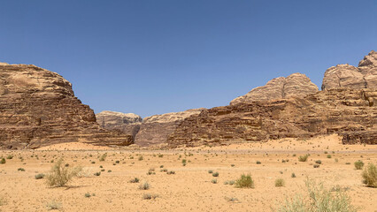 Vast, sunny desert landscape in Wadi Rum, Jordan, featuring towering sandstone mountains and a clear blue sky over the arid, sandy terrain.
