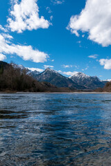 Fototapeta premium Clear Lech River reflecting snowy mountains and clouds near Füssen, Bavaria, Germany – tranquil alpine landscape with blue sky and crystal water