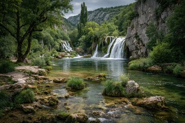 Obraz premium Tranquil waterfall cascading into a clear river, surrounded by lush greenery and rocky terrain
