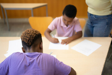 Young students sitting at desk and writing, with a teacher standing nearby supervising