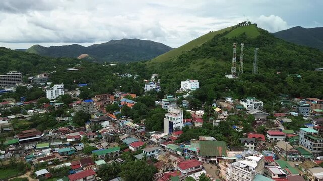 Daytime aerial partial orbit above Coron on Busuanga Island, showing Mount Tapyas, telecom towers and dense town rooftops set among lush green hills under a clear blue sky.