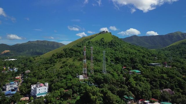 Aerial view toward Mount Tapyas over Coron with telecom towers on the lower hill green forested slopes nearby buildings and a bright daylight mountain backdrop
