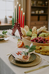 Kwanzaa table setting featuring kinara with seven lit candles, fruit basket with bananas, grapes, apples, corn, and place setting with decorative napkin and greeting card