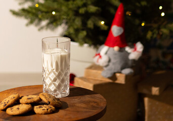 Milk and cookies on wooden board near Christmas tree in soft light. Boy in pajamas prepares sweet treats and drink for Santa before bedtime. Cozy winter holiday atmosphere.