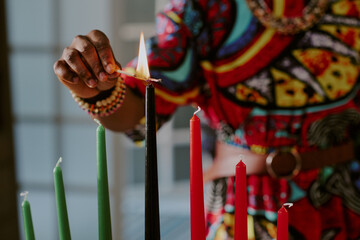 Black woman lighting black candle on kinara with matchstick during Kwanzaa celebration, colorful patterned clothing and beaded bracelets visible, focus on hand and candles