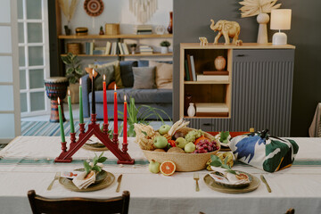 Kwanzaa table setting featuring kinara with seven candles, fruit basket centerpiece, wrapped gift and two place settings arranged for celebration in modern living room